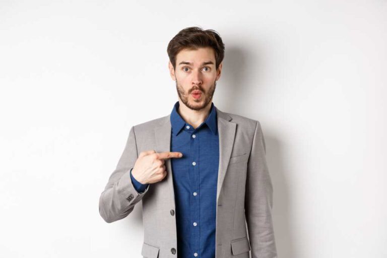 Who me. Surprised man in suit pointing at himself with disbelief, looking confused at camera, standing on white background